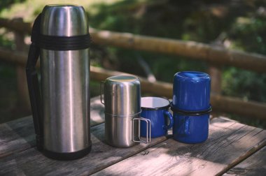 Dışarıdaki ahşap bir masada mavi emaye kupalarının yanında çelik bir termos duruyor. Rahat bir kamp ortamının mükemmel bir tasviri ya da doğada sıradan bir kahve molası..