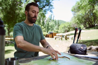 Bir adam doğa manzaralı, güneşli bir parkta piknik masasında yürüyüş haritası üzerinde çalışıyor. Etrafı yürüyüş direkleri, bir çanta ve doğal yeşilliklerle çevrili..