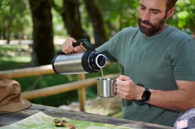 Bir adam dışarıdayken termostan sıcak bir sıvıyı bardağa boşaltıyor. Sahnede kırsal bir masa ve orman yabanı var. Rahatlama ve keşif çağrıştırıyor..