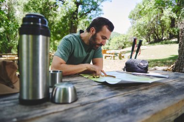 Parkta güneşli bir günde tahta bir masada oturan bir adam yürüyüş ve kamp ekipmanlarıyla çevrili bir şekilde haritaya bakıyor. Yürüyüşünü planlıyor..