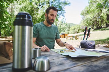 Bir birey piknik masasında doğal bir ortamda yürüyüş için haritayı inceliyor. Olay yerinde yürüyüş ekipmanları, termal matara ve yemyeşil şeyler var..