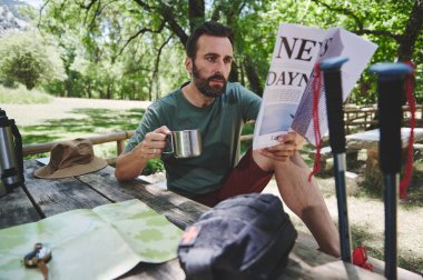 Rahat bir adam, güneşli bir günde sakin bir orman parkında yürüyüş yaparken kahve içerken gazete okur..