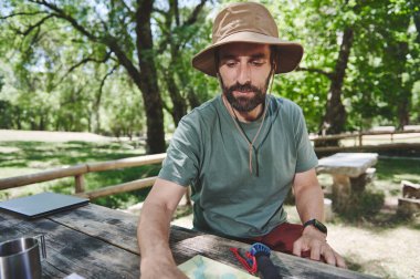 Giyim kuşamında bir piknik masasında oturmuş haritayı inceliyor ve sakin bir açık hava deneyiminin tadını çıkarıyor..