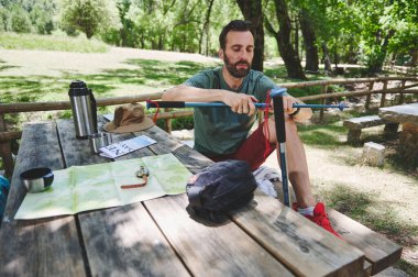 Tahta bir piknik masasında yürüyüş teçhizatı ve haritalarla çevrili yürüyüş direklerini toplayan adam, huzurlu bir orman ortamının tadını çıkarıyor, açık hava aktivitelerini ve rahatlamayı temsil ediyor..