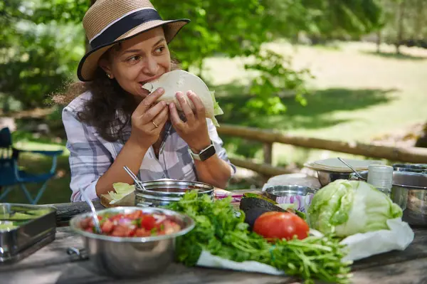 Bir kadın taze sebzelerle dolu elle hazırlanmış bir tortilladan zevk alır, doğal bir ortamda, ahşap bir masada salata ve domates gibi malzemelerle çevrili bir yerde oturur..