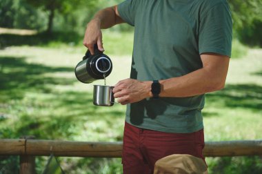 Bir adam, serin ve güneşli bir ortamda termostan metal bir bardağa içki doldurur. Macerayı, rahatlamayı sembolize eder ve sakin bir kamp deneyimi sırasında doğayla yeniden bağlantı kurar..