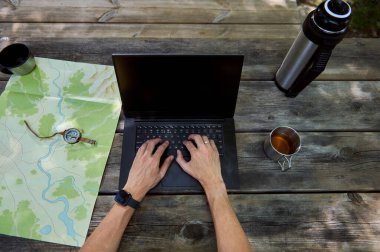 Dışarıdaki çalışma alanını bir dizüstü bilgisayar, harita, pusula ve kahveyle tasvir eden resim. Uzaktan çalışma, macera planlaması ve huzurlu bir doğal ortamda verimli çoklu görev sunar.