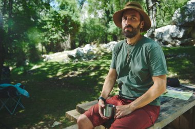 Sakallı bir adam ormanın içindeki tahta bir masada oturuyor. Sahne, güneşli bir yaz gününde macera ve sükunetin vurgulandığı eğlenceli bir kamp deneyimi olduğunu gösteriyor..