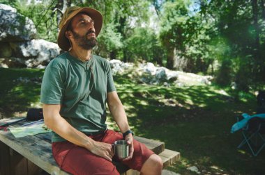 Sakallı bir adam, günlük kıyafetlerini giymiş, bir orman kamp alanındaki ahşap bankta dinleniyor, elinde metal bir kupa tutuyor, güneş ışığı ve huzurlu doğal manzarayla çevrili..