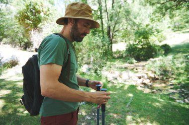 Yürüyüş yapan bir adam yürüyüş direklerini düzeltirken açık havada yemyeşil bir ortamda sırt çantası ve yürüyüş şapkasıyla.