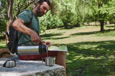 Açık havada genç bir adam termostan bir bardağa içecek dolduruyor. Günlük giysiler, bir sırt çantası giyiyor ve bir haritayı inceliyor, keşiflerinin tadını çıkarıyor..