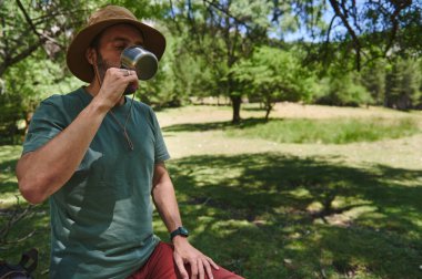 Uzun ağaçlarla çevrili yeşil, güneşli bir alanda oturmuş, kamp ve yürüyüş deneyimini kucaklarken, şapka takan bir adam, metal bardaktan su içmekten zevk alır..