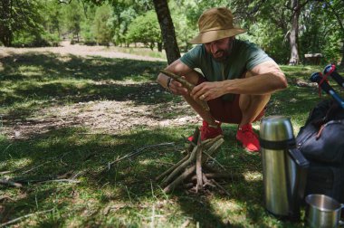 Bir adam, yemyeşil bir ormanda kamp yaparken kamp ateşi yakmak için odun topluyor. Açık hava macerası ve doğa ruhunu somutlaştıran güneşli doğada kamp malzemeleriyle çevrili..