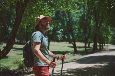 Açık bir yaz gününde ormanda yürüyüş yapan, şapka takan ve yürüyüş direkleri tutan bir adam. Etrafı toprak bir patika boyunca canlı yeşil ağaçlarla çevrili..