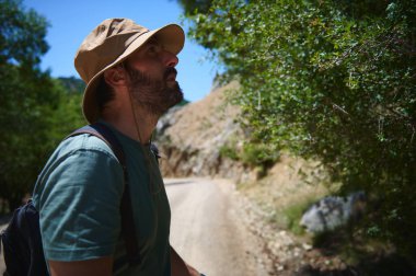 Güneşli bir günde şapka takan bir adam dağ manzarası ve yeşilliğin tadını çıkarıyor, aktif ve maceraperest bir yaşam tarzını doğanın ortasında somutlaştırıyor..