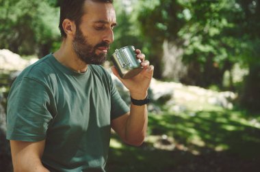 Yeşil gömlekli bir adam, doğal bir çevrede ağaçlar ve yeşilliklerle birlikte metalik bir bardaktan su içer..