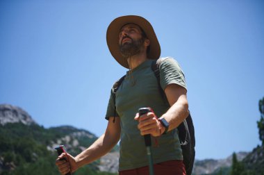 Yürüyüş şapkası ve ekipmanıyla güneşli bir günde verimli bir vadide gezinen bir adam macera ve açık hava keşifleri sergiliyor..