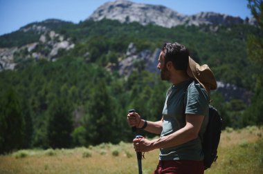 Yeşil dağların arasında doğa güzelliğinin tadını çıkaran bir yürüyüşçü yürüyüş direkleri taşıyor ve sakin bir açık havada korunmak için şapka takıyor..