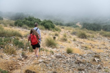 Bir çift yürüyüşçü sisli havada kayalık bir dağ yolundan geçer, keşifleri, doğa dalgıçlığını ve doğal ortamdaki macerayı vurgular..