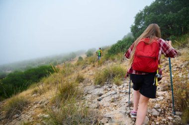 Yürüyüşçüler yeşil bitki örtüsüyle çevrili sisli ve kayalık bir dağ yolundan geçerek keşfi, zindeliği ve doğayla bağlantıyı vurguluyorlar..