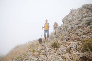 Bir köpek eşliğinde bir çift yürüyüşçü sis kaplı bir çevrede kayalık bir yamaçtan inerek macerayı, keşfi ve doğayla bağlantıyı somutlaştırıyor..