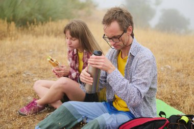 Bir adam ve bir kadın yürüyüş sırasında çimenlerde oturur, kuru arazi manzarası ile çevrili bir yemeği paylaşır ve dışarıda dinlenmenin tadını çıkarırlar..