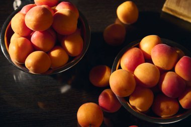 Koyu ahşap bir yüzeye karşı metalik kaselerde sergilenen olgun, canlı kayısılardan oluşan bir koleksiyon. Işıklandırma onların doğal renklerini güçlendiriyor, davetkar ve taze bir ortam yaratıyor..