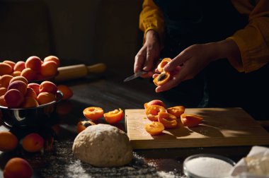 Bir insan taze meyve, un ve hamur ile çevrili bir kesme tahtasındaki bıçakla kayısıları dilimler ve sıcak, kırsal bir mutfak ortamı yaratır..