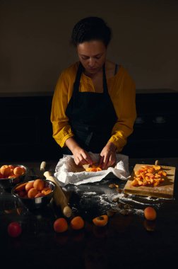 Sarı bluzlu ve siyah önlüklü bir kadın en az mutfakta kayısılı börek hazırlıyor. Kayısılar, hamurlar ve çatal bıçaklar koyu bir tezgahta düzgünce düzenlenir..