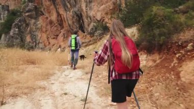 İki genç insan ve köpekleri aktif ve sağlıklı bir yaşam tarzını kucaklıyor, engebeli dağ patikalarında yürüyüş yapıyorlar. Yürüyüş direkleri ve sırt çantalarıyla açık hava macerasını ve doğanın güzelliğini seviyorlar..