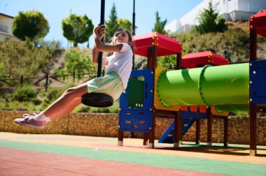 Neşeli çocuk renkli bir parkta salıncakta sallanırken eğleniyor. Parlak, enerjik ve bir yaz gününün neşesini yakalıyor..