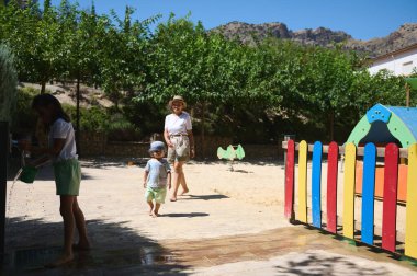 Neşeli yapılar ve parlak güneş ışığı altında neşeli çevreler arasında bir grup çocuğun eşlik ettiği manzaralı bir oyun alanı..