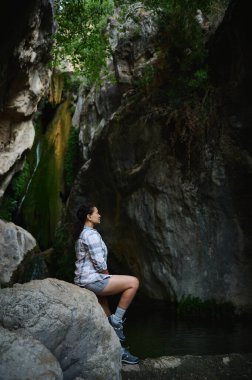 Kayaların ve yeşilliklerin arasında sakin bir derenin kenarında oturan bir kadının huzurlu bir anı. Doğal ortam sükunet ve doğa ile bağlantı yayar..