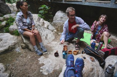 Üç arkadaş açık havada derenin kenarındaki kayalarda oturup kahvenin tadını çıkarıyorlar. Kamp malzemeleri ve yemek malzemeleri olay yerine katkıda bulunuyor, yoldaşlık ve doğal keşif temaları çağrıştırıyor.
