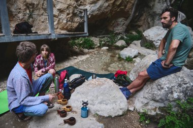 Arkadaşlar dışarıda toplandı, bir köprünün yanındaki kayaların üzerinde oturdular, doğayla çevrili rahat bir anın tadını çıkardılar. Kamp malzemeleri ve bir köpek sakin sahneyi tamamlıyor..