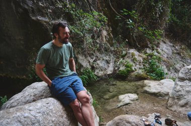 Gülümseyen bir adam, yeşil bitki örtüsü ve doğal manzaralarla çevrili, berrak bir derenin yakınındaki bir kayanın üzerinde oturuyor. Huzurun tadını çıkarıyor..