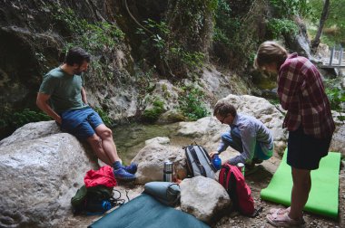Arkadaşlar yeşil yaprakların arasında kayalık bir dere kenarında toplanarak sırt çantaları ve malzemelerle kamp yapmaya hazırlanıyorlar. Doğanın güzelliği ve yoldaşlığı için sakin bir inziva..