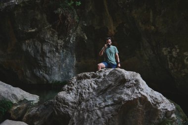 Bir adam, doğal kaya oluşumları ve yemyeşil yemyeşil yemyeşil olan sakin bir kanyonla çevrili bir kayanın üzerinde dururken sıcak bir içecekten zevk alır. Sahne huzur ve macera taşır.