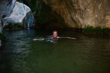 Huzurlu, kristal berrak doğal havuzda yüzen bir adamın görüntüsü yemyeşil bitki örtüsü ve yükselen kaya oluşumlarının arasına yerleşmiş, barış ve doğal bir bağ hissi uyandırıyor..