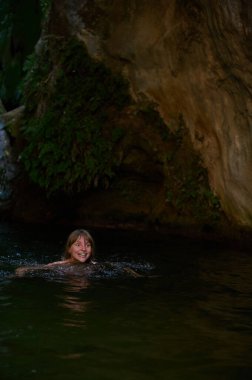 Gülümseyen bir kadının neşeli bir sahnesi, kayalık uçurumlara ve yemyeşil bitki örtüsüne dayanan, huzurlu bir doğal su bedenine daldırılmış, mutluluğu ve doğayla bağlantıyı yakalayan,.