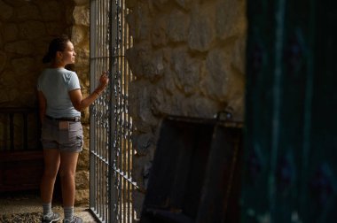 Yıpranmış bir duvarın yanında duran bir kadın, sıcak, kırsal tonlar ve gölgelerle çevrili dekoratif bir demir pencereden dışarı bakar, sakin ve yansıtıcı bir ortam izlenimi verir..