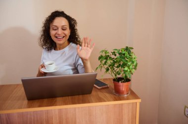Evde dizüstü bilgisayar kullanan gülümseyen bir kadın, video konferansından zevk alıyor. Elinde bir kahve fincanı tutarak, coşkuyla el sallıyor, ahşap bir masada oturmuş, atmosferi güçlendiriyor..