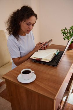 Bir ajandaya danışırken bilgisayarını ve telefonunu kullanarak masa başına odaklanmış bir kadın. Bir kahve ve bitki ile rahat çalışma alanı ev ortamında verimli bir çalışma ortamı sağlamaktadır..