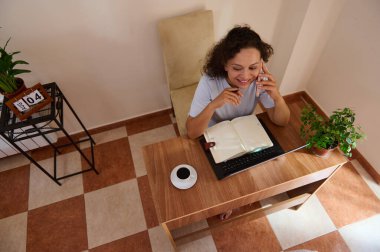 Profesyonel bir kadın evde uzaktan çalışıyor, dizüstü bilgisayar kullanırken telefonla konuşuyor. Çalışma alanı bitkiler ve şık bir ahşap masa içeriyor..