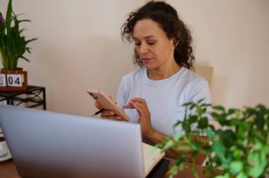 Freelancer ev atmosferinde mobil bir cihaz ve dizüstü bilgisayar kullanarak görevlere odaklanıyor, uzaktan çalışmayı vurguluyor.