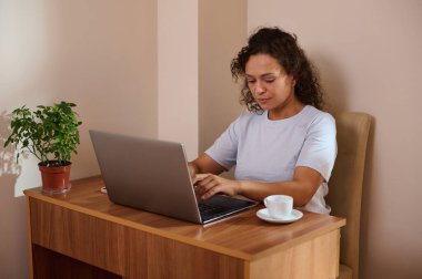 Uzaktan kumandalı bir kadın, rahat bir ev ortamında dizüstü bilgisayarda, bir bitki ve kahve fincanı ile birlikte, ortamı geliştiren bir masa başında oturuyor..