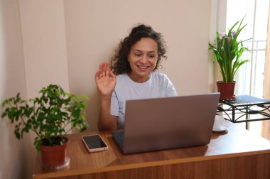 Gülümseyen bir kadın, rahat, bitki süslemeli bir ev ortamında dizüstü bilgisayarında uzaktan çalışma ve iletişim gösteren sanal bir buluşmaya katılıyor. Profesyonellik ve rahatlık hissi veriyor..