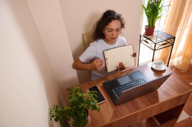 Uzaktan çalışan bir kadın, ahşap bir masada dizüstü bilgisayar ve defter kullanarak doğal ışıklandırma ve ev bitkileriyle hoş bir ofis ortamı yaratıyor..