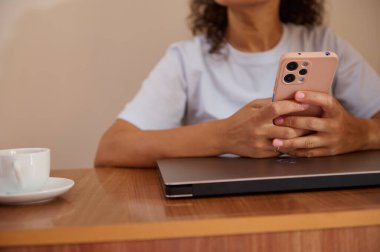 Bir kadın, ev ortamında rahat bir masada otururken, telekomünikasyon ve rahatlamayı gösteren bir akıllı telefonu tutarak birden fazla iş yaparken görülüyor..