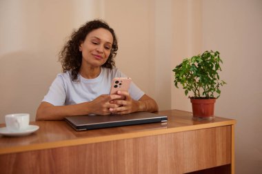 Bir kadın akıllı telefonuna odaklanmış bir şekilde masada oturuyor. Kapalı bir dizüstü bilgisayar, saksı bitkisi ve kahve fincanı modern uzaktan kumandalı sıcak ve üretken bir atmosfer yaratır..